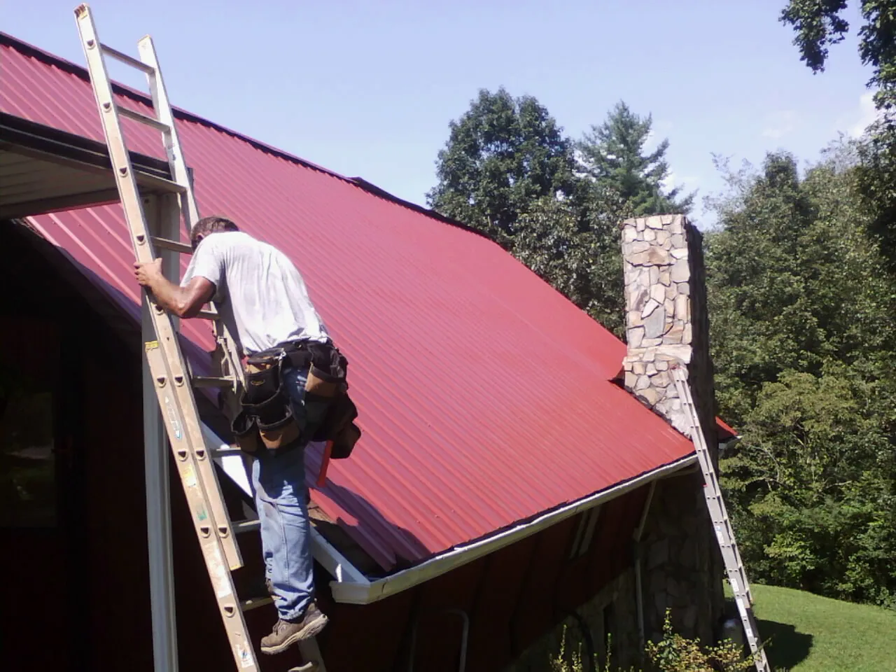 Roofer on a ladder beside a red metal roof and stone chimney — Soffit & Fascia Repair by Roofing Brothers in Canajoharie