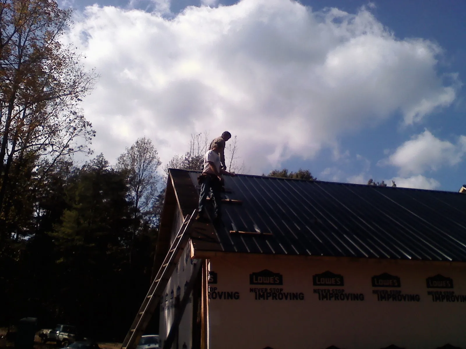 Roofer fastening dark metal panels high against a cloudy sky, from a Roof Inspection job in Canajoharie